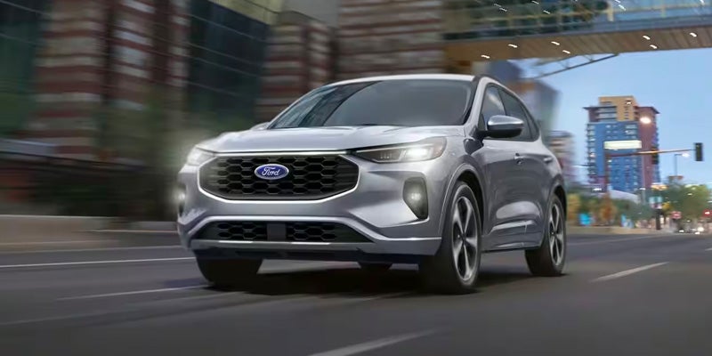 silver 2024 ford escape plug-in hybrid with headlights on driving on a street a city skyline in the background